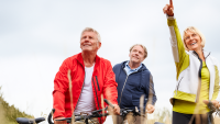Tre seniorer på cykler i naturen smiler og peger.