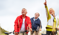 Tre seniorer på cykler i naturen smiler og peger.